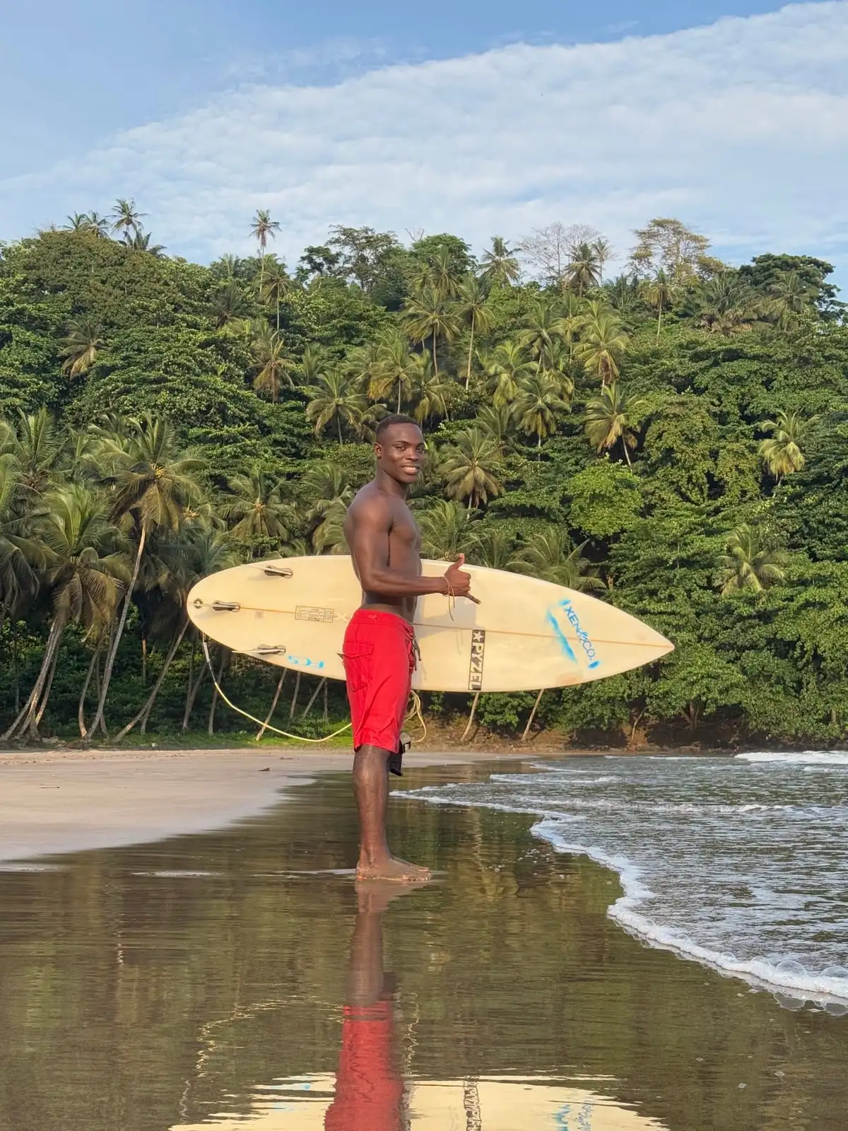 A surfer in red shorts stands on wet sand holding a surfboard, with palm-covered hills behind and a reflection in the shallow water.