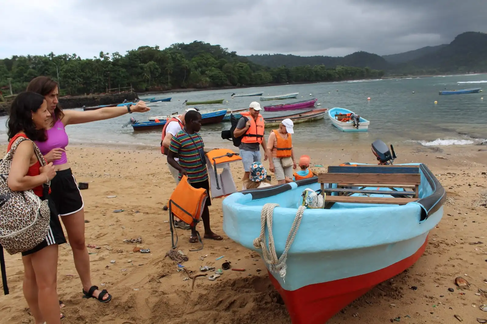 People in life jackets gather around a light-blue boat on a sandy beach while one of them points across the bay.