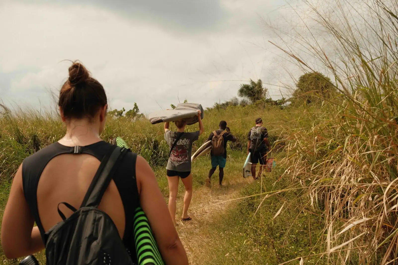 A line of people carry surfboards and bags along a grassy path toward the sea.