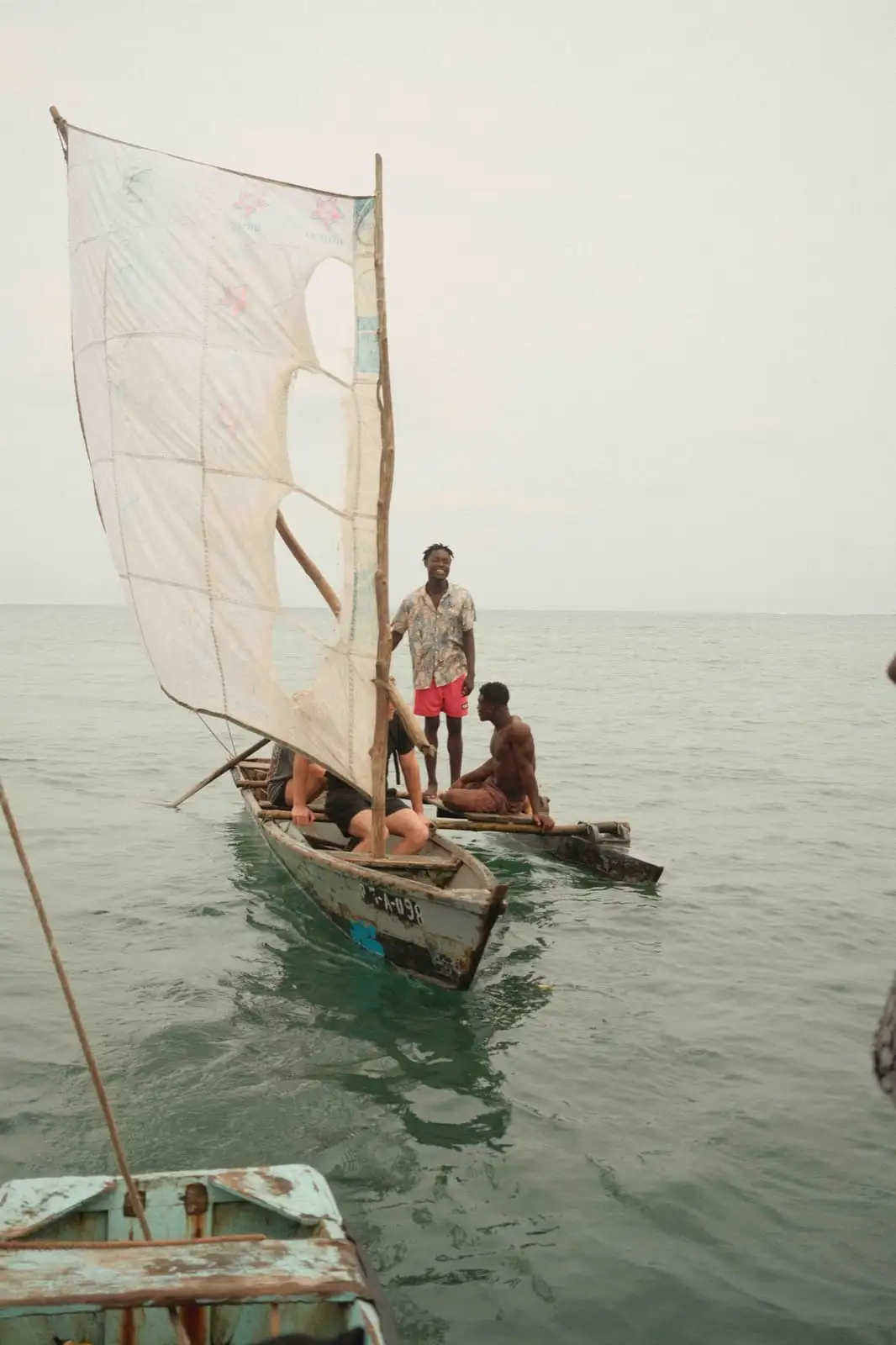 A small wooden boat with a patched white sail carries several people across calm green water.