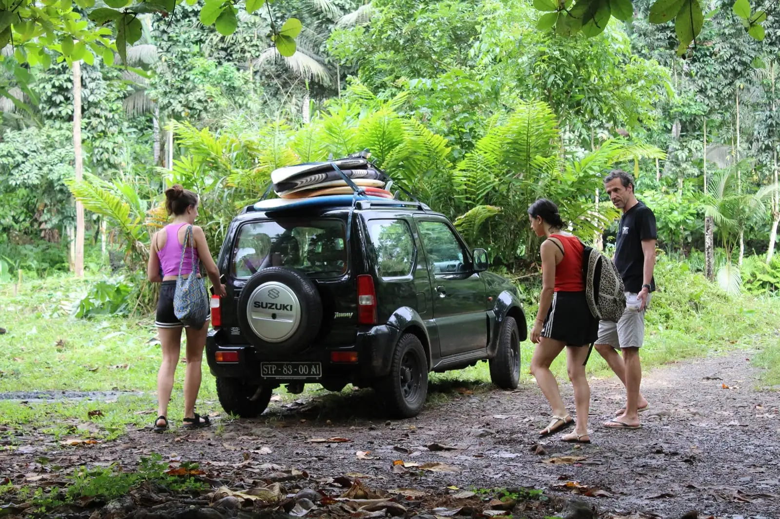 A dark green jeep with boards on its roof parked on a forest track, with people walking past.