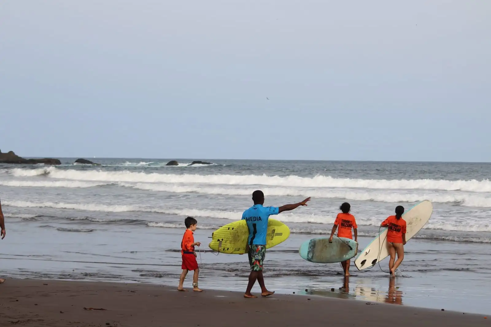 An adult points out to sea while several younger people carrying surfboards walk along the beach under an overcast sky.