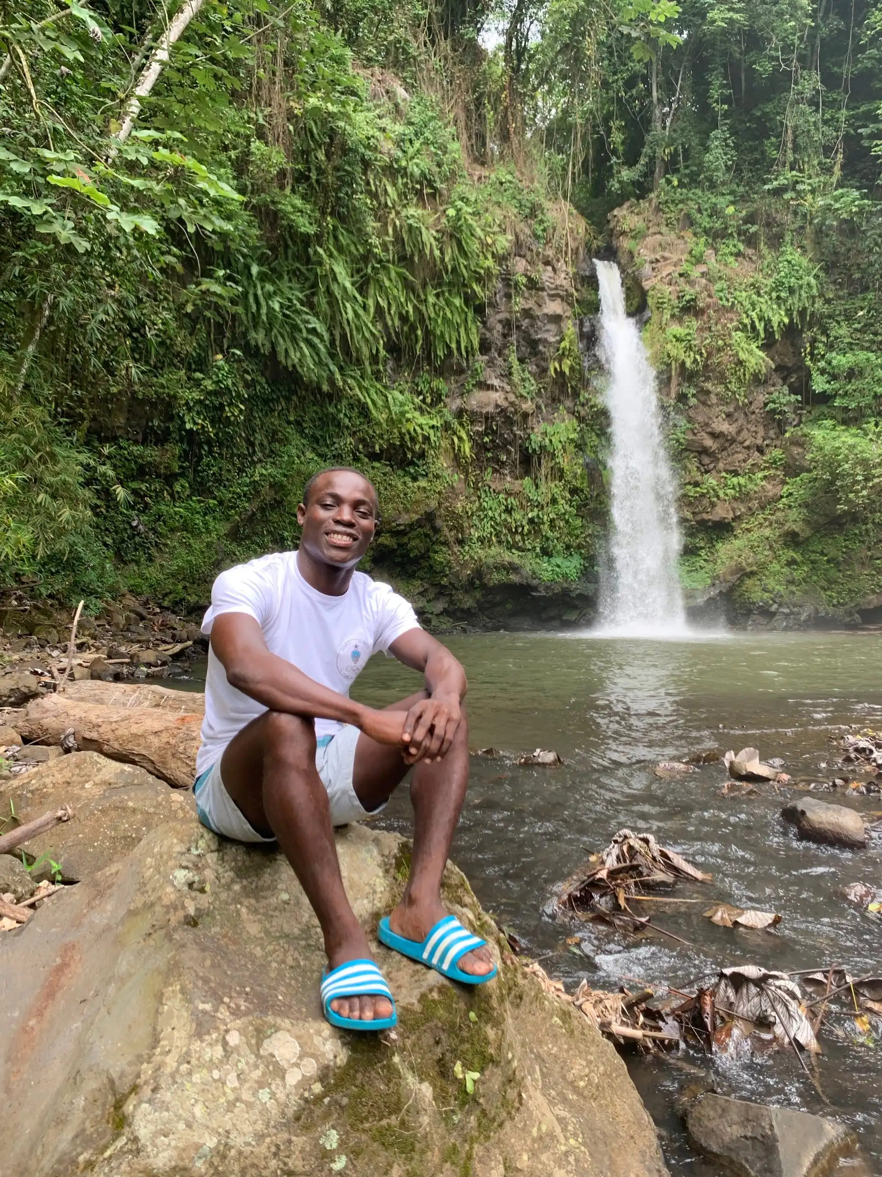 A smiling man in a white t-shirt and shorts sits on a rock beside a river, with a waterfall and green forest behind him.