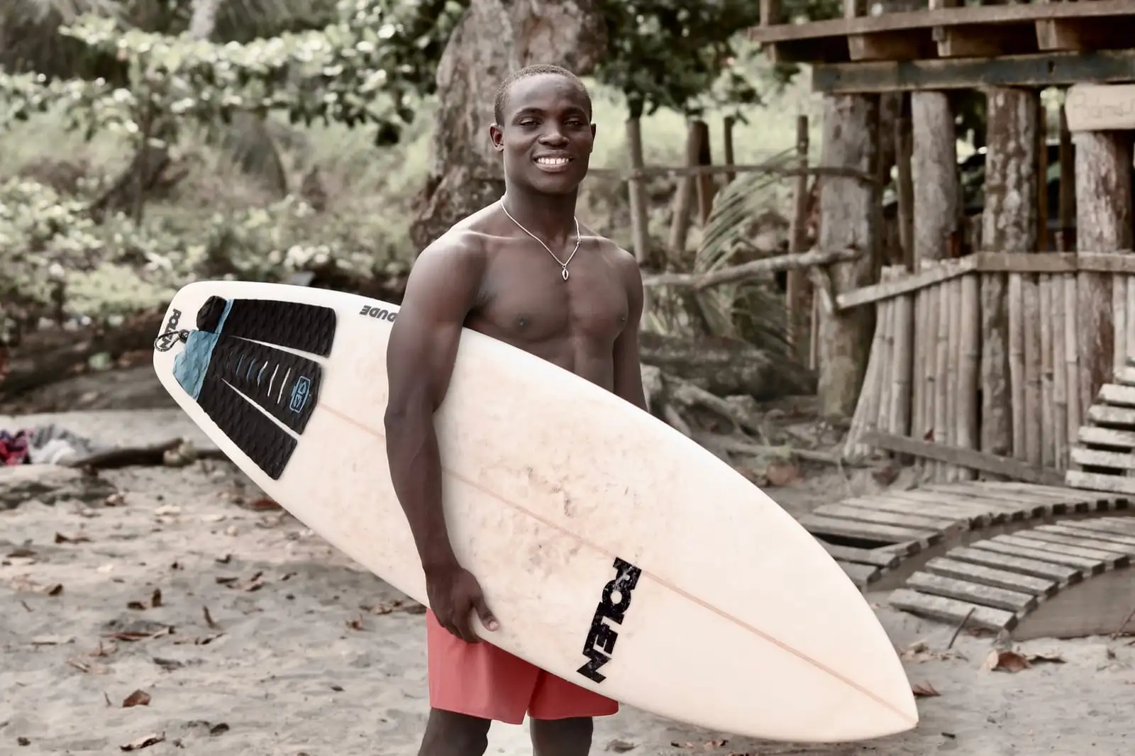 A smiling surfer holds a white surfboard under one arm on a sandy beach, with wooden structures behind.