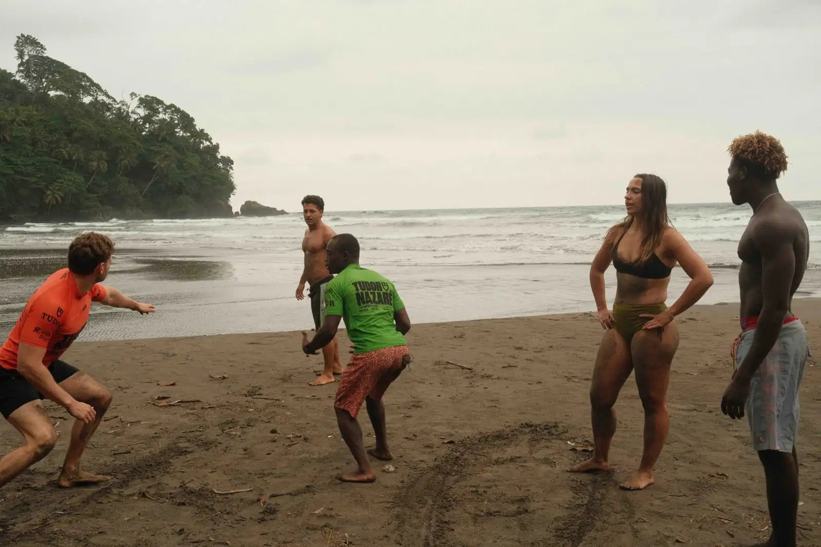 A small group of people in athletic poses on a dark-sand beach, with a forested headland in the background.