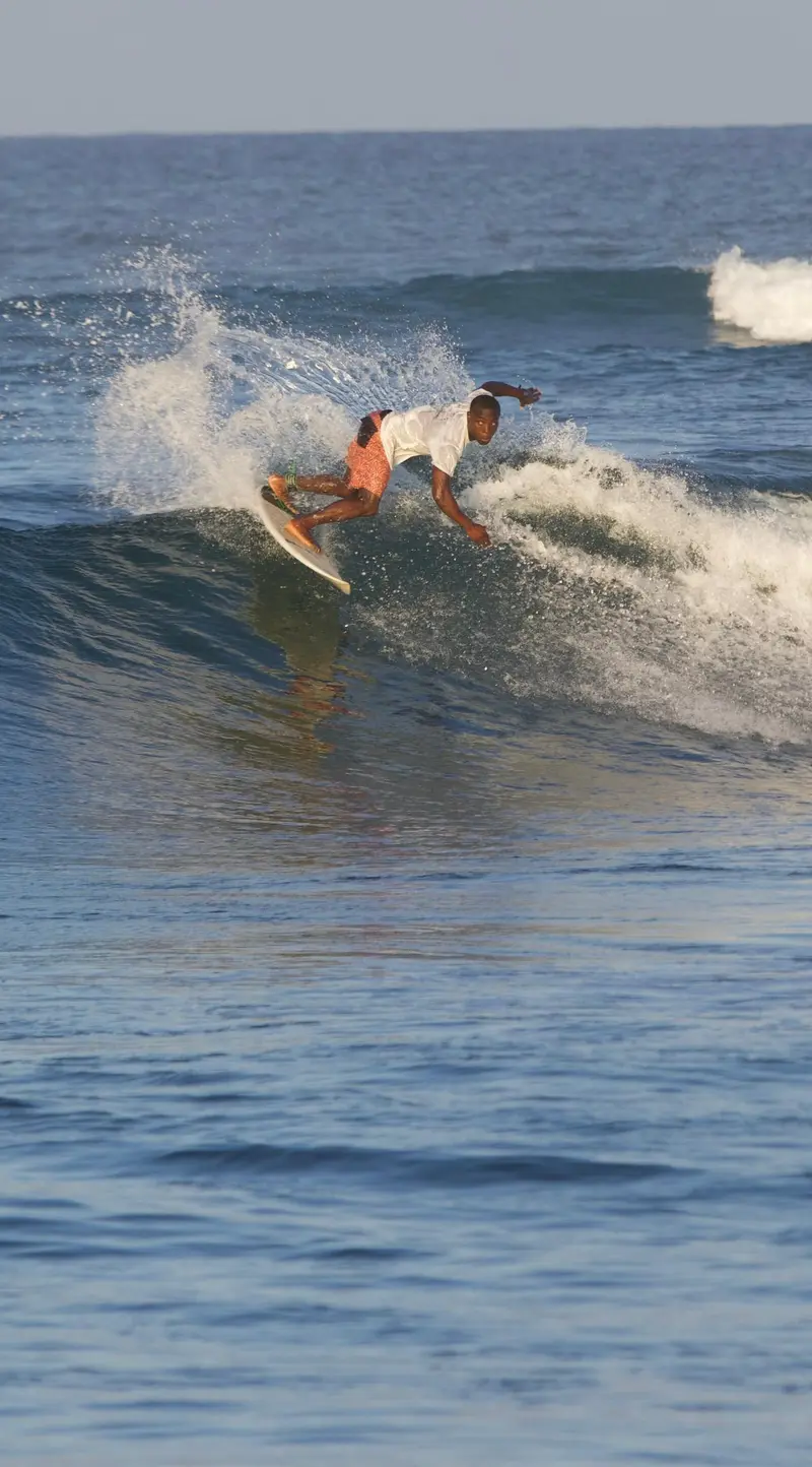 A surfer in a white t-shirt and orange shorts leans into a turn on a breaking wave, throwing up white spray in warm afternoon light.